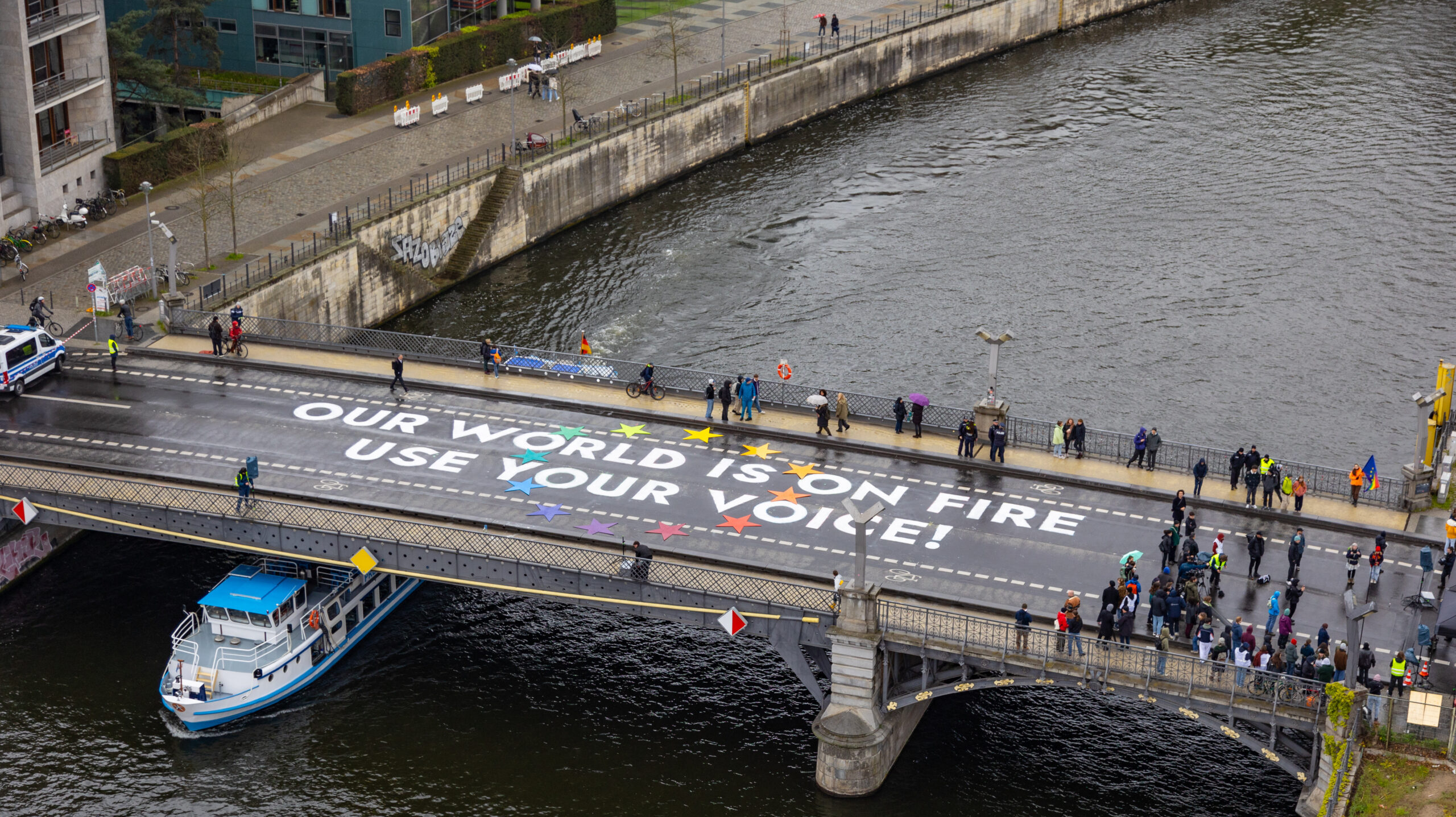 Aktion am 19.04.2024: Schriftzug auf der Marschallbrücke in Berlin: "Our world is on fire - use your voice" umgeben von zwölf bunten Sternen